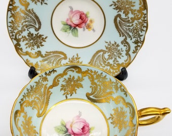 May include: A vintage teacup and saucer set with a delicate floral design and gold accents. The cup and saucer are a light blue color with a white center featuring a pink rose and gold trim.