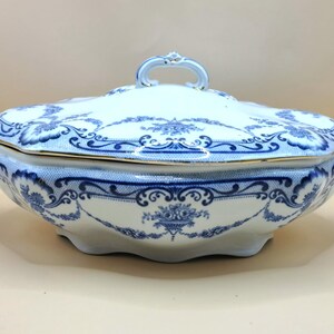 May include: A white and blue porcelain serving dish with a lid. The dish features a blue floral pattern and gold trim. It has handles on the sides and a handle on the lid. The dish is oval-shaped and sits on a light-colored surface.