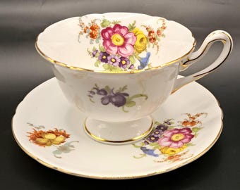 May include: A white teacup and saucer set with a floral pattern in shades of pink, yellow, purple, and blue. The cup has a scalloped rim and a gold trim. The saucer has a matching floral design.