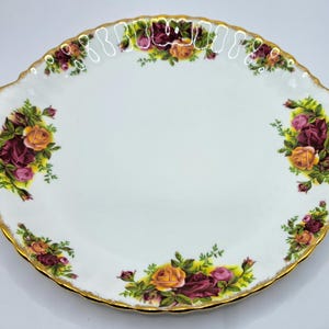 Puede incluir: Plato ovalado blanco con borde dorado y estampado floral de rosas. Las rosas son en tonos amarillo, rosa y burdeos. El plato tiene un borde festoneado.
