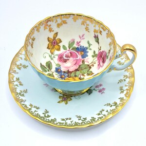 Könnte beinhalten: Ein Vintage-Tee-Set, bestehend aus einer Tasse und einer Untertasse. Die Tasse und die Untertasse sind hellblau mit goldenen Verzierungen. Die Innenseite der Tasse zeigt ein Blumenmuster mit rosa Rosen, blauen Blumen und gelben Akzenten.
