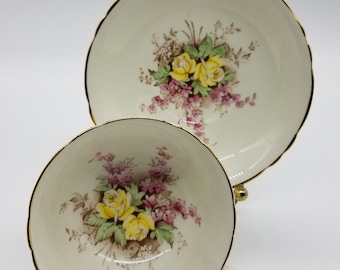 May include: A vintage teacup and saucer set with a floral design featuring yellow roses and pink flowers. The set has a white background with gold trim.