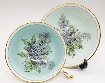 May include: A light blue teacup and saucer set with a floral design of purple and white lilac flowers and green leaves. The set has a gold trim around the edges.