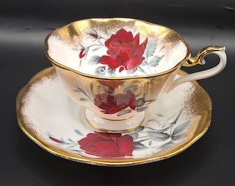 May include: A white teacup and saucer set with a gold rim and red rose floral pattern. The teacup has a delicate handle and the saucer has a scalloped edge.
