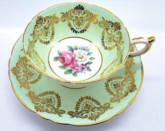 Peut inclure: Une tasse à thé et une soucoupe en vert avec des bordures dorées et un motif floral. La tasse et la soucoupe ont un bord festonné délicat.