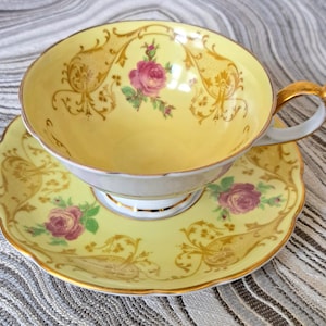 May include: A vintage teacup and saucer set. The cup and saucer are yellow with gold filigree and pink rose floral designs. The cup has a gold handle and trim. The saucer has a scalloped edge.