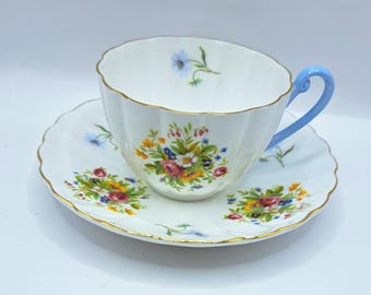 Puede incluir: Un juego de taza y platillo blancos con un diseño floral. La taza tiene un asa azul claro y un borde festoneado. El platillo y la taza están decorados con flores coloridas y ribetes dorados.