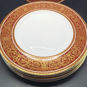 May include: A set of four white dinner plates with a red and gold decorative border. The border features a floral pattern with gold accents.