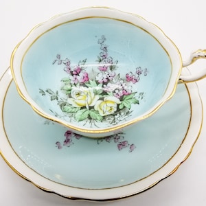 May include: A light blue teacup and saucer set with a floral design featuring yellow roses and pink flowers. The set has a gold trim around the edges.
