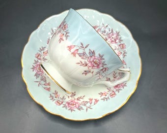 Può includere: Un set vintage di tazza e piattino da tè. La tazza e il piattino sono azzurri con motivi floreali rosa e bordi dorati. La tazza è inclinata sul piattino, mostrando il design delicato.
