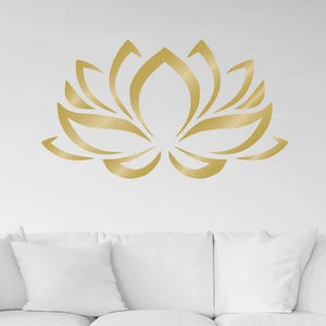 May include: A gold-coloured lotus flower wall decal featuring a stylised, symmetrical design with pointed petals. The graphic is applied to a plain white wall positioned directly above a white sofa and two wooden coffee tables in a room.