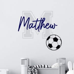 Könnte beinhalten: Ein Wandaufkleber mit einem großen hellgrauen Buchstaben M, über dem der Name Matthew in dunkelblauer Schreibschrift steht, ergänzt durch eine schwarz-weiße Fußballgrafik, die sich unten rechts neben dem Buchstaben befindet.