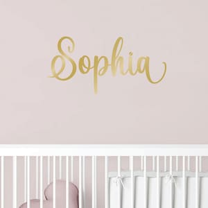Könnte beinhalten: Ein goldfarbener Wandaufkleber mit dem Namen Sophia in einer geschwungenen Schreibschrift. Der Schriftzug ist auf einer hellrosa Wand angebracht, direkt über einem weißen Kinderbett mit einem herzförmigen Kissen und weißer Bettwäsche.