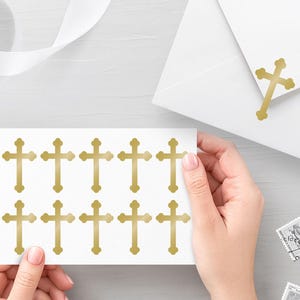 Może przedstawiać: Biały prostokątny arkusz z dziesięcioma złotymi naklejkami w kształcie krzyża, ułożonymi w dwóch rzędach po pięć sztuk. Pasująca złota naklejka w kształcie krzyża znajduje się na klapce białej koperty. Krzyże mają metaliczne wykończenie.