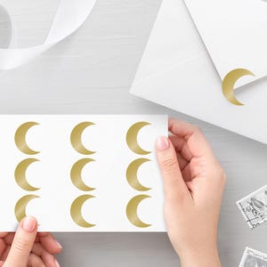 Könnte beinhalten: Ein weißes rechteckiges Blatt mit neun goldenen Halbmond-Aufklebern, die in drei Spalten und drei Reihen angeordnet sind. Ein einzelner goldener Halbmond-Aufkleber ist zudem auf der Klappe eines weißen Briefumschlags angebracht.