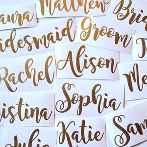 Peut inclure: Une collection d'étiquettes adhésives rectangulaires blanches individuelles présentant des noms et des titres en écriture cursive dorée métallique. Le texte inclut Laura, Matthew, Bride, Bridesmaid, Groom, Tom, John, Rachel, Alison, Megan, Sophia, Christina, Katie et Samantha.