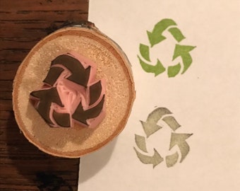 Könnte beinhalten: Ein Holzstempel mit dem Recycling-Symbol, das in den Gummi geschnitzt ist. Der Stempel wird verwendet, um grüne und graue Abdrücke des Symbols auf weißem Papier zu erzeugen. Der Stempel besteht aus einem Querschnitt eines Astes.
