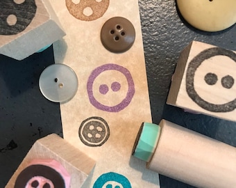 May include: An assortment of buttons and button-themed stamps. The buttons vary in size, color, and material, including a large beige button and a small, clear button. The stamps feature button designs in various colors, such as brown, purple, and teal.