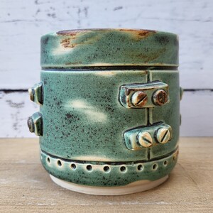 May include: A green ceramic mug with a distressed finish and metal accents. The mug has a white rim and a row of small holes around the bottom.