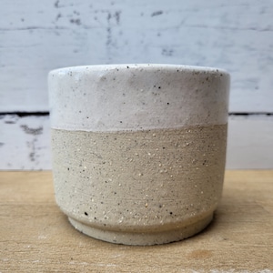 May include: A white and beige speckled ceramic planter with a rounded bottom. The planter is made of a rough, textured material.