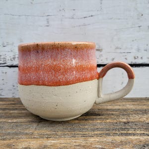 May include: A handcrafted ceramic mug with a rounded base and a large handle. The mug has a speckled, off-white base, transitioning to an orange band and a brown rim. The mug is displayed on a wooden surface.