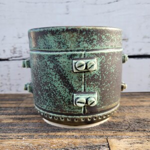 May include: A green and brown ceramic mug with a speckled glaze and a decorative band of metal accents. The mug has two sets of screws on the front and sides.