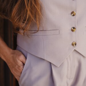 May include: A light purple pantsuit with a vest and matching pants. The vest has a pointed collar and gold buttons. The pants have a front pleat and a pocket on the side.