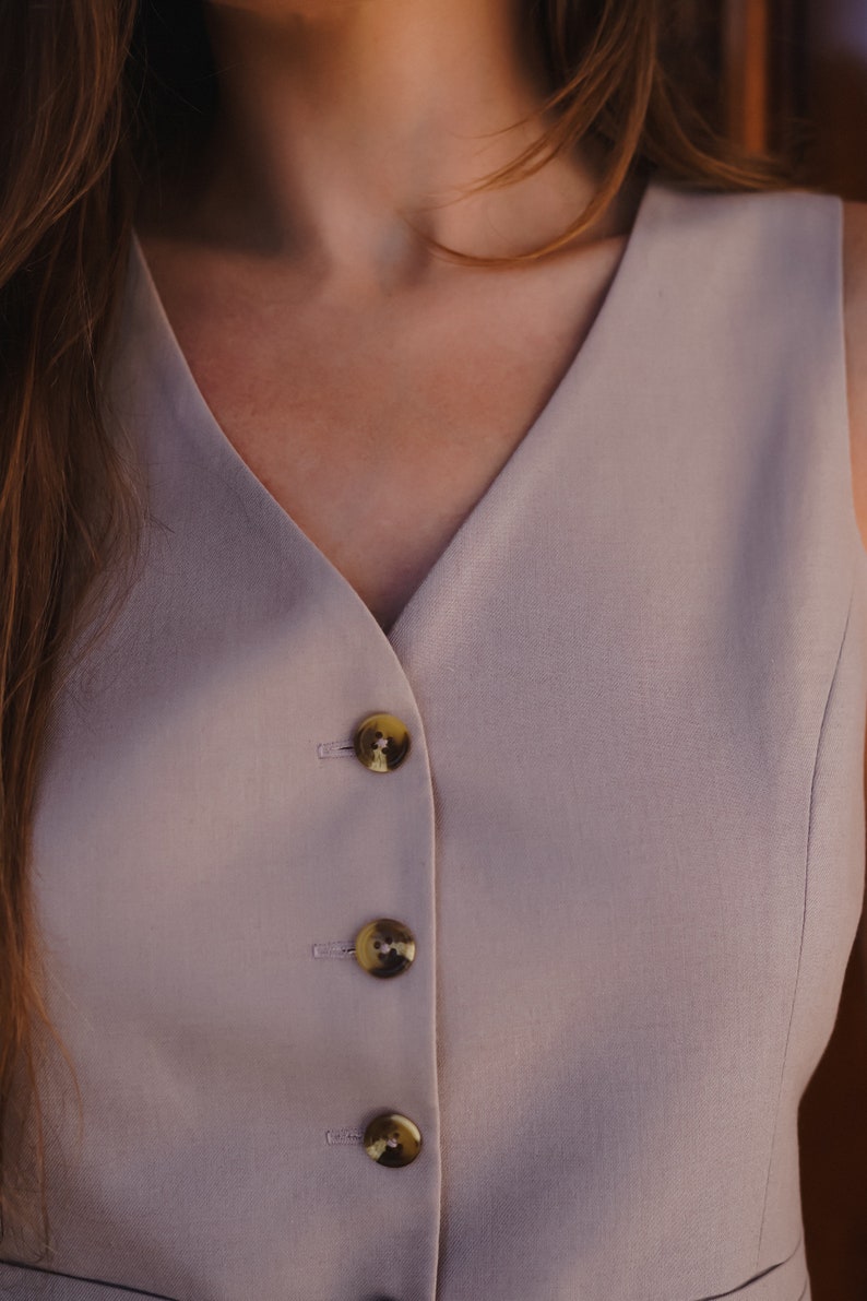 May include: A light beige sleeveless top with a V-neckline and three gold buttons down the front.