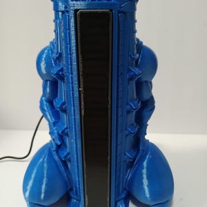 May include: A blue 3D-printed object with a rectangular black panel in the center. The object has a cylindrical shape with rounded protrusions and a textured surface. A black cord extends from the base.