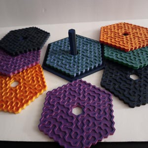 Könnte beinhalten: Eine Sammlung farbenfroher, sechseckiger Untersetzer in Orange, Lila, Grün und Marineblau. Jeder Untersetzer hat ein strukturiertes, wellenartiges Muster und ein zentrales Loch. Ein blauer sechseckiger Sockel mit einem zentralen Pfosten ist ebenfalls vorhanden.