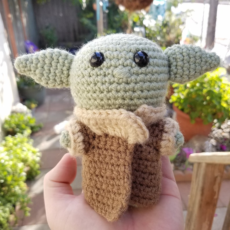 May include: A crocheted green and brown plush toy resembling a character from the Star Wars franchise. The toy has large black eyes and a brown crocheted coat.