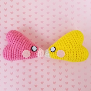 May include: Two crocheted heart-shaped plushies, one pink and one yellow, with black eyes and pink noses, facing each other on a pink background with a pattern of small pink hearts.