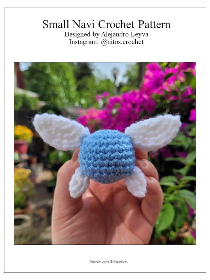 May include: A small blue crocheted figure with white wings. The figure is being held in a hand. The background is blurry and shows a garden with pink flowers.