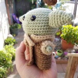 May include: A crocheted green and brown plush toy resembling a character from the Star Wars franchise. The toy has large black eyes and a pointed ear.