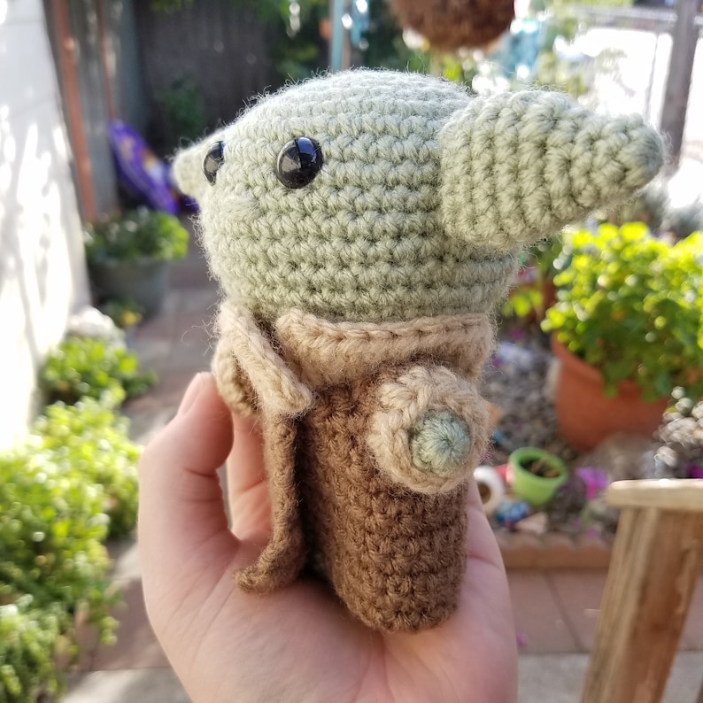 May include: A crocheted green and brown plush toy resembling a character from the Star Wars franchise. The toy has large black eyes and a pointed ear.