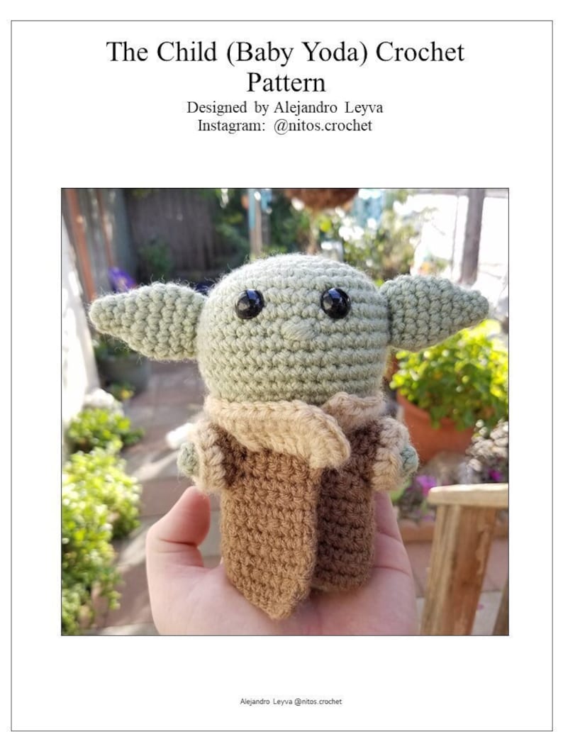 May include: A crocheted Baby Yoda doll, also known as The Child, wearing a brown robe. The doll is green with large ears and black eyes. The doll is being held in a hand.