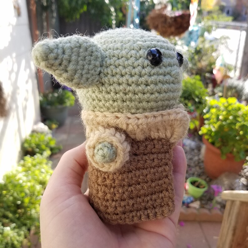 May include: A crocheted green and brown plush toy with large black eyes. The toy is shaped like a small creature with a large head and small body.