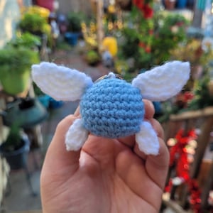 May include: A crocheted blue and white winged creature keychain. The body is a light blue sphere with white wings and feet. The keychain is held in a hand against a blurred background of greenery.