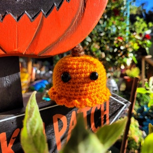 May include: A handmade, crocheted orange pumpkin with black button eyes and a brown stem. The pumpkin is sitting near a sign that says "KID PATCH" in orange letters. The sign is black with an orange pumpkin on top.