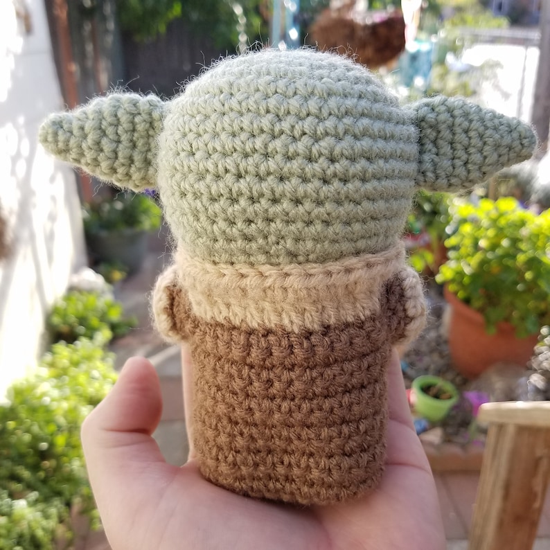 May include: A crocheted green and brown plush toy resembling a character from the Star Wars franchise. The toy is being held in a person's hand.