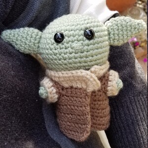 May include: A crocheted green and brown plush toy resembling a character from the Star Wars franchise. The toy has large black eyes and a brown crocheted coat.