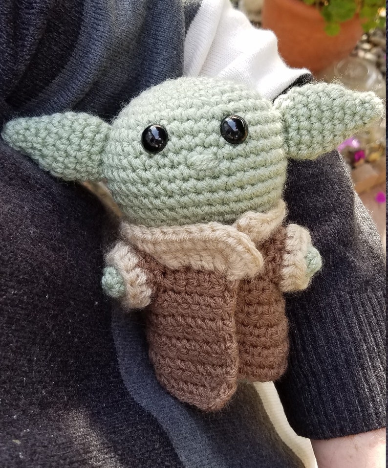 May include: A crocheted green and brown plush toy resembling a character from the Star Wars franchise. The toy has large black eyes and a brown crocheted coat.