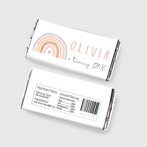 Op de afbeelding: Een afdrukbare sjabloon voor een chocoladeverpakking met een regenboog in de kleuren roze, oranje en grijs. De tekst "Olivia wordt EEN" staat op de verpakking. De verpakking is ontworpen voor een eerste verjaardag.