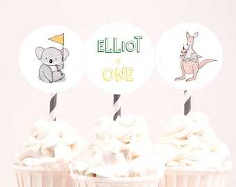 Puede incluir: Tres adornos para cupcakes con fondo blanco. Los adornos representan un koala sosteniendo una bandera, un canguro sosteniendo un cupcake y un círculo con el texto "Elliot is One".