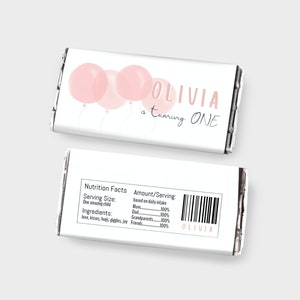 May include: A printable chocolate bar wrapper template featuring pink watercolour balloons and the text "Olivia is turning ONE". The back of the wrapper has a nutrition facts label with a barcode and the name "Olivia".
