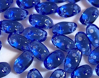 Puede incluir: Una colección de pequeñas cuentas de vidrio ovaladas de color azul vibrante. Las cuentas tienen un acabado ligeramente iridiscente y dos pequeños agujeros para enhebrar.