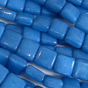 May include: A collection of bright blue square-shaped beads with a smooth, glossy finish. The beads are arranged in multiple rows, showcasing their uniform shape and vibrant color. Each bead features rounded edges and a flat surface, reflecting light.