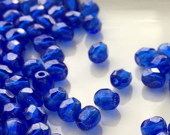 May include: A large quantity of small, faceted, cobalt blue glass beads. The beads are scattered on a white surface.