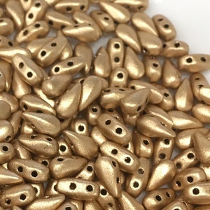 May include: A collection of metallic gold teardrop-shaped beads. Each bead has two holes for stringing. The beads have a matte finish and are scattered across the image, suggesting a craft supply or jewelry component.