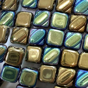 May include: An assortment of square beads in shades of gold, teal, and blue. The beads have a metallic, iridescent finish, and some feature diagonal grooves. The beads are arranged closely together, filling the frame.